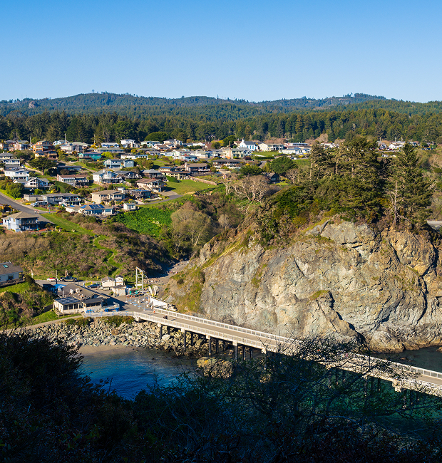 Scenic view of large rock outcrop and bridge over water with small town and mountains in background.