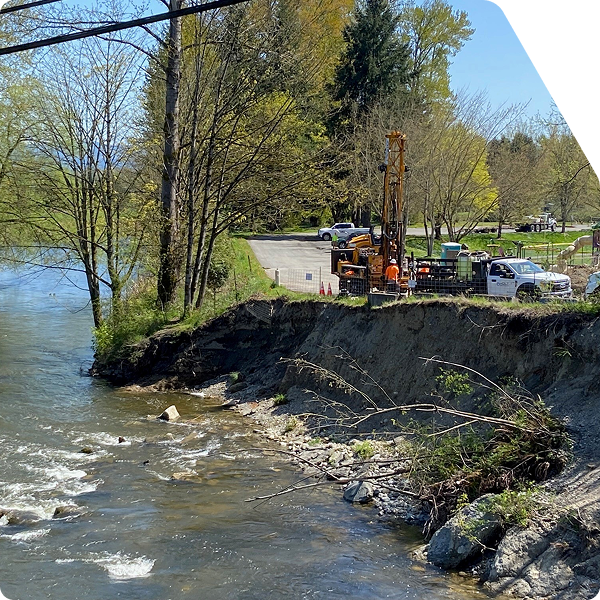 Geotechnical sampling with drill rig near slope along riverbank.