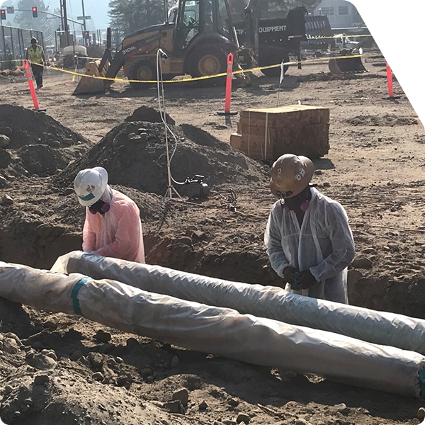 Two workers in hazardous material suits in front of a wrapped pipe.
